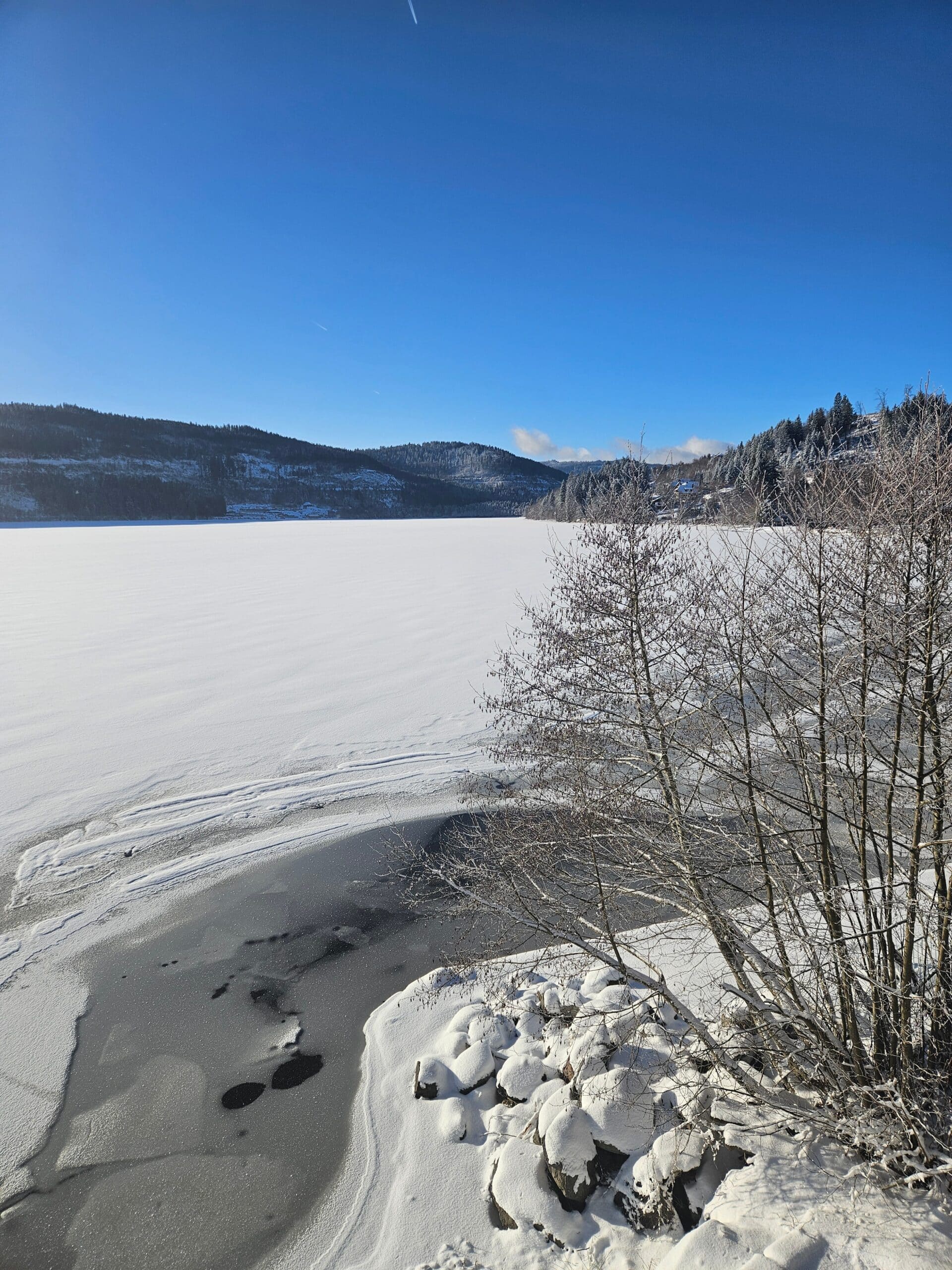 Verschneiter, teilweise zugefrorener Titisee im Hochschwarzwald im Winter bei klarem blauem Himmel, mit bewaldeten Hügeln im Hintergrund und schneebedeckten Ufersteinen im Vordergrund.
