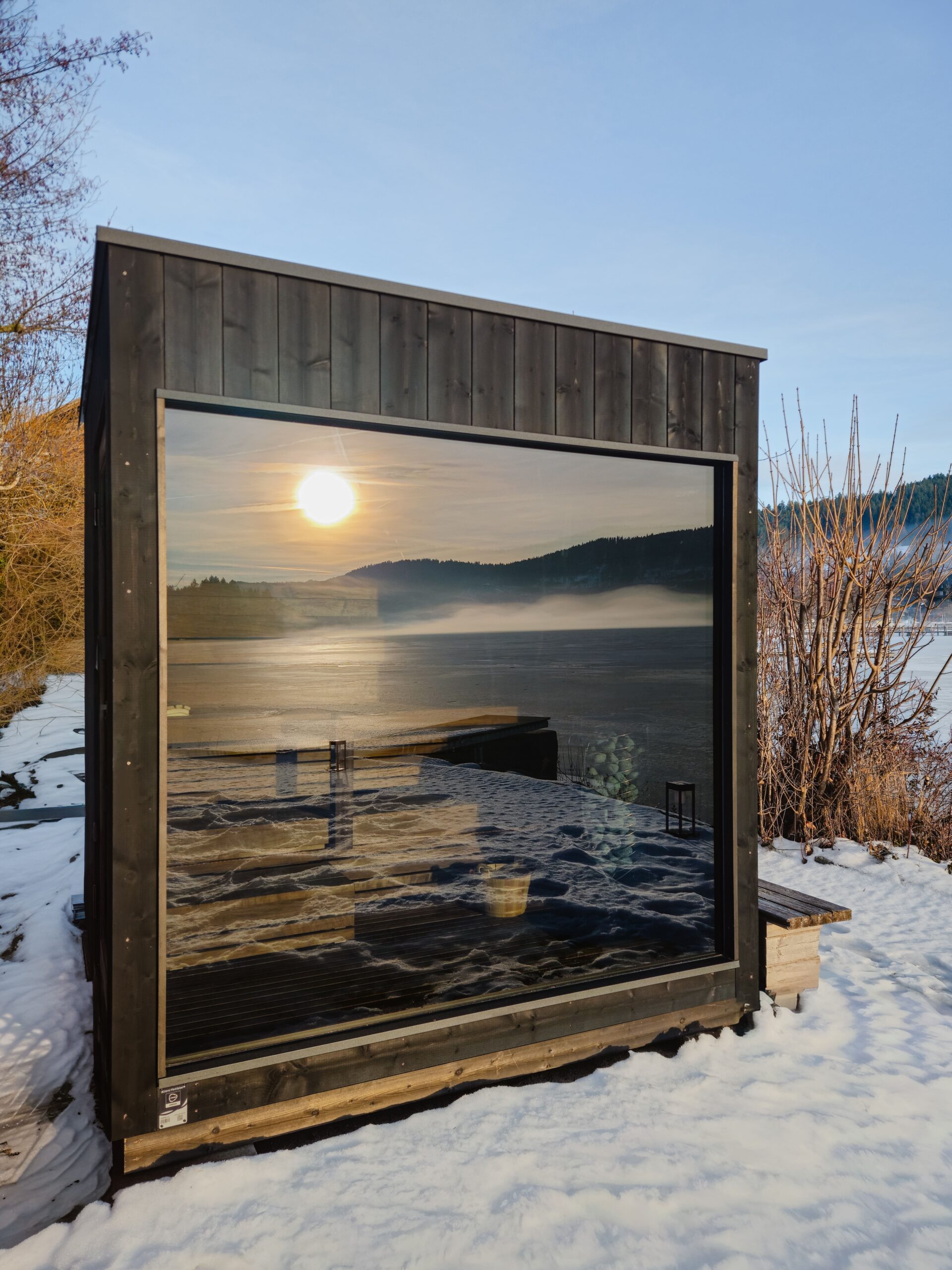 Winter-See-Sauna am verschneiten Seeufer, dunkle Holzfassade mit großer Glasfront, Spiegelung von Sonne, Eis und ruhiger Winterlandschaft.