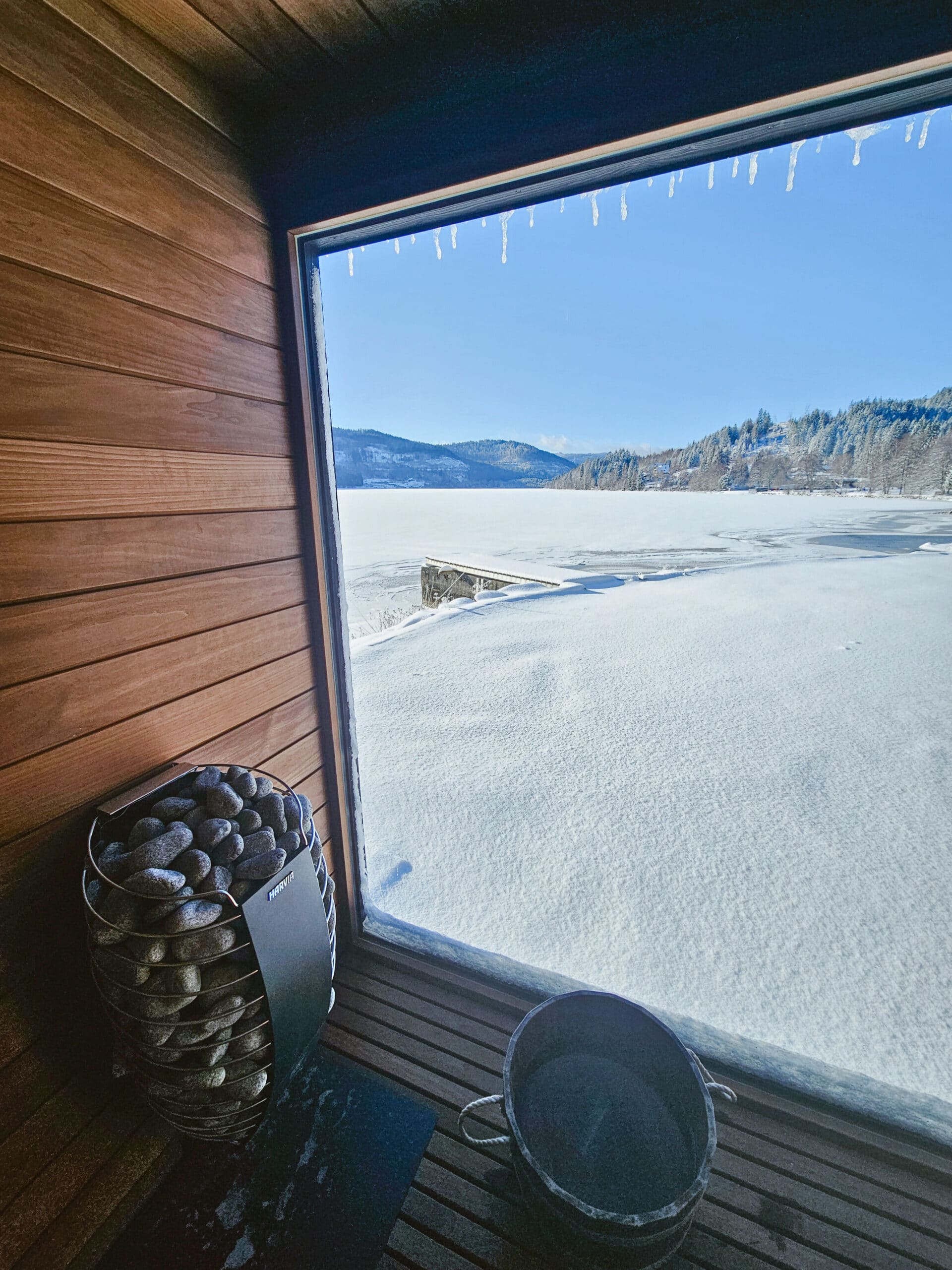 Blick aus einer holzverkleideten Pop-up Sauna am Strand des Seehotel Wiesler durch ein großes Fenster auf den verschneiten, zugefrorenen Titisee mit Winterlandschaft und klarem blauem Himmel. Links steht ein Saunaofen mit Steinen, am Boden ein Holz-Wasserbottich.